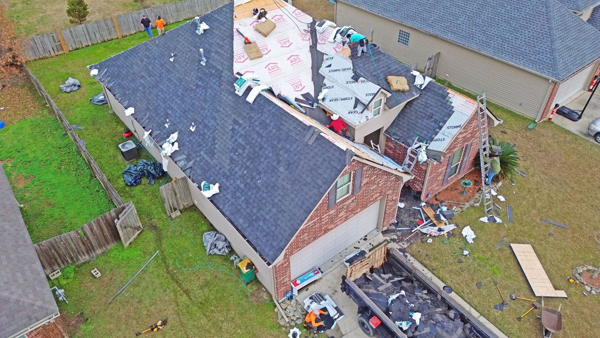 Does Insurance Cover a New Roof?Aerial view of a residential roof replacement in progress, showing workers on the roof, construction materials, and a ladder. The area around the house is filled with discarded roofing materials and tools, highlighting an active roofing project in Gulfport, MS. This is an example of how insurance pays for roof replacement.